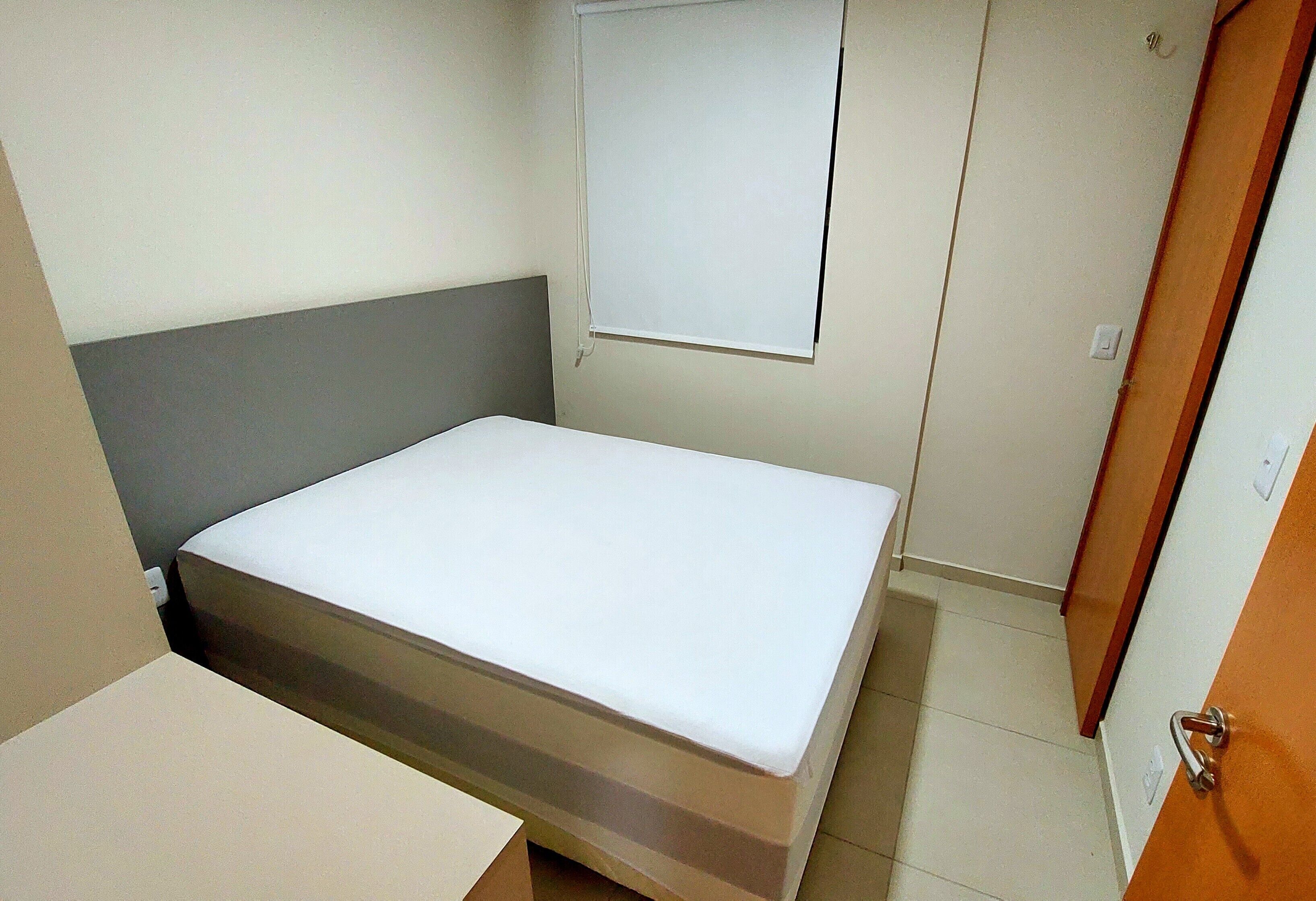 A small, single-bed room with a grey headboard, a white mattress, and a window with a white blind.