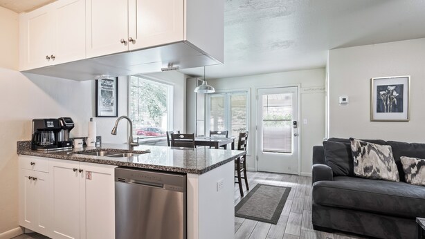 A modern kitchen with white cabinets, a built-in dishwasher, and a granite countertop. There is a dining area with a gray sofa and a coffee machine. - Newly Renovated Unit: Contemporary Interiors, Minutes from Fine Dining & Wold Class Skiing! (Park City)