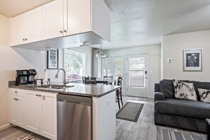 Una cocina moderna con gabinetes blancos, lavavajillas integrado y encimera de granito. Cuenta con una zona de comedor con sofá gris y cafetera.