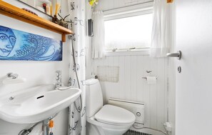 Et badeværelse med et hvidt håndvask, toilet og brusebad. Der er et vindue med udsigt, og et billede på væggen.