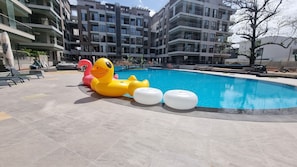 Una piscina con juguetes inflables, un suelo de baldosas y un edificio de varios pisos al fondo.