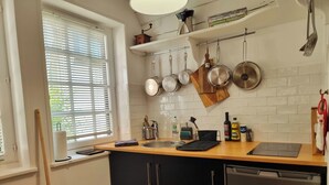 Una cocina con encimera de madera, un fregadero y armarios. Hay ollas colgadas en la pared, una ventana con persianas y una cuchara de madera apoyada contra la pared.