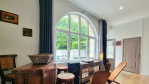 A room with a large arched window, a wooden table with a basket, a chair, and a cabinet with drawers.