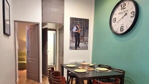 A dining area with a table set for two, a clock on the wall, and a door leading to another room.