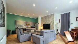 A modern living room with grey sofas, a coffee table, and a TV. A kitchen with grey cabinets and a stone wall is visible in the background.