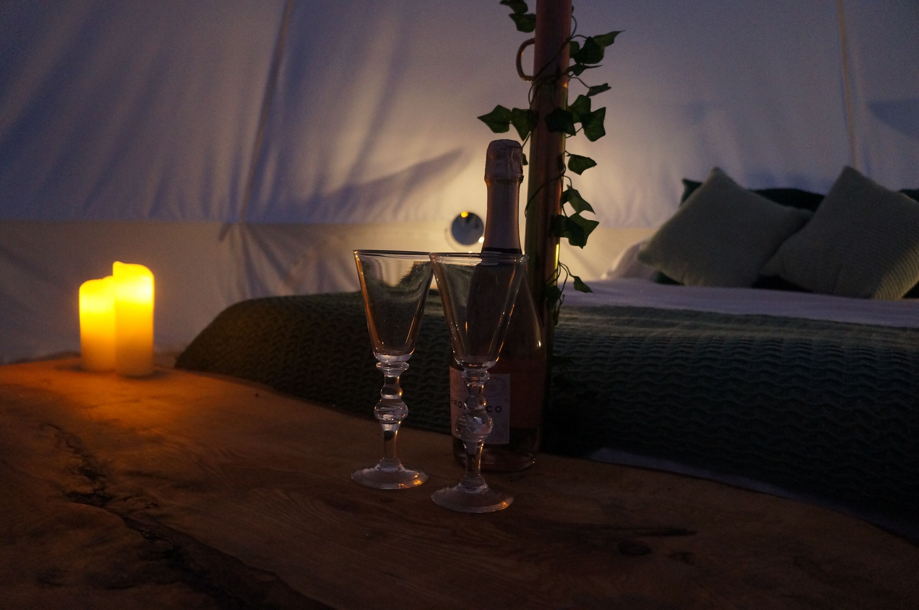 A wooden table with two wine glasses, a bottle, and a lit candle in a tent-like room with a bed and pillows.
