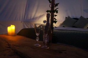 A wooden table with two wine glasses, a bottle, and a lit candle in a tent-like room with a bed and pillows. - The Bells Glamping (Morpeth)