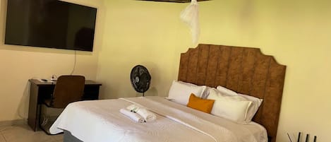 A hotel room with a bed, a desk, a chair, a fan, and a TV.