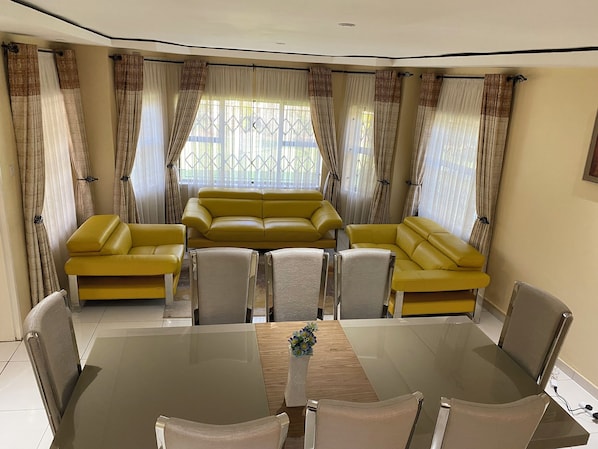 A living room with a yellow sofa set, a glass dining table, and a decorative vase. - Goshen Creek Guesthouse  (Harare)