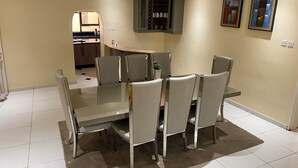 Een eetgedeelte met een glazen tafel en comfortabele stoelen, een keuken op de achtergrond, en twee ingelijste kunstwerken aan de muur.