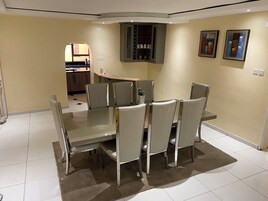 Un comedor con una mesa de vidrio y sillas acolchadas, una cocina al fondo y dos cuadros en la pared.