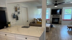 Een moderne keuken met een marmeren aanrecht en witte kasten, een woonehoek met een bank en open haard, en een eethoek met een tafel en stoelen.