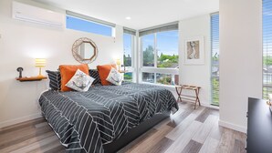 A modern bedroom with a large bed, a bedside table with a lamp, a window with blinds, and a view of greenery outside.