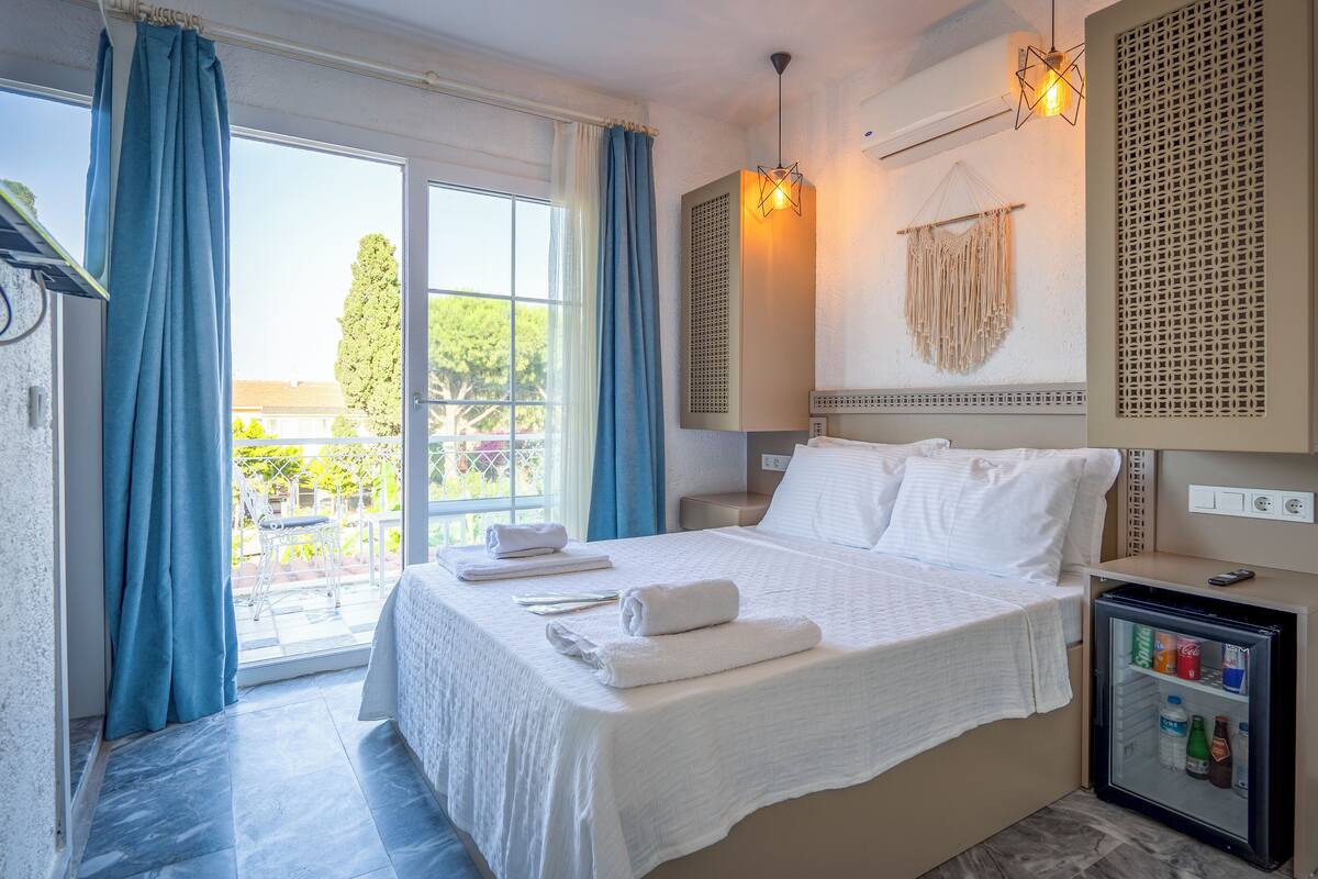 Une chambre d’hôtel avec un lit, un petit réfrigérateur, une fenêtre avec des rideaux et une vue sur la verdure environnante.