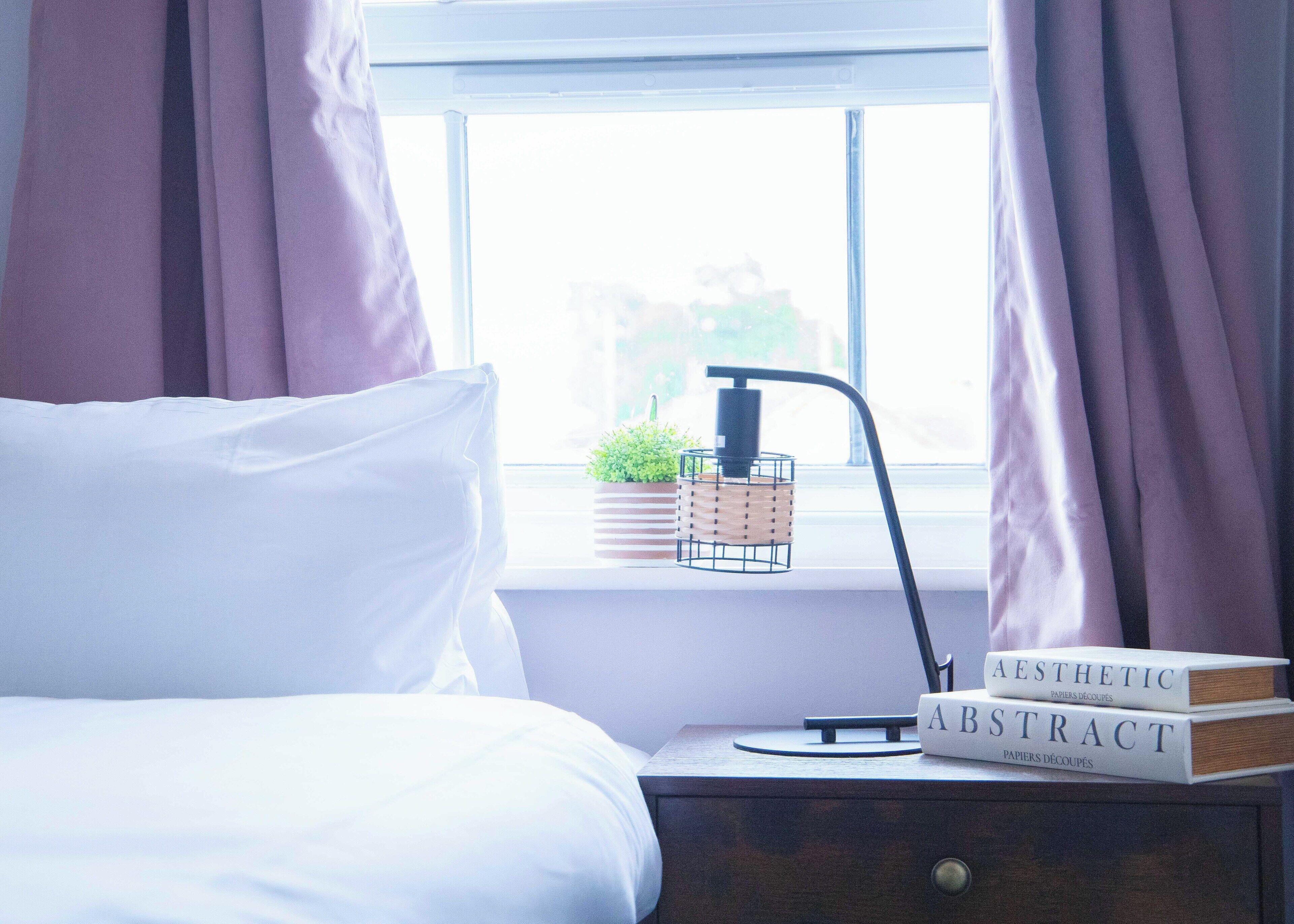 En pænt redt seng med hvide sengetøj, et natbord med en lampe, en lille potteplante og to bøger med titlerne 'AESTHETIC' og 'ABSTRACT'.
