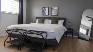 A bedroom with a bed, a bedside table, a nightstand, a mirror, and a window with blinds. - Modern home 3-BR Retreat — Quiet, Cozy, & Family-Friendly in Fresno. (Fresno)