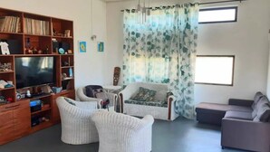 Ein Wohnzimmer mit Fernseher, Bücherregal, zwei weißen Sesseln, einem grauen Sofa und einem Fenster mit Vorhang.