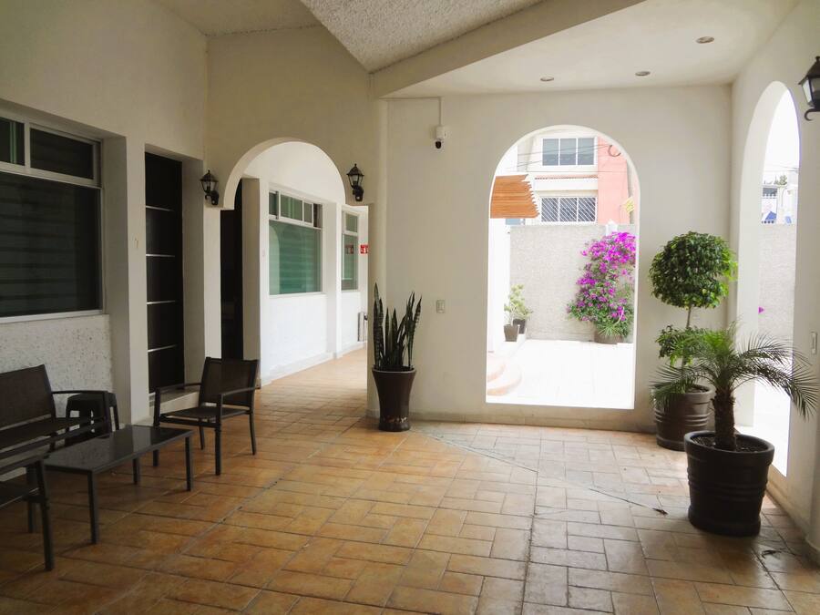 A corridor with tiled flooring, benches, potted plants, and arched doorways leading to a courtyard with a view of a building and a tree.