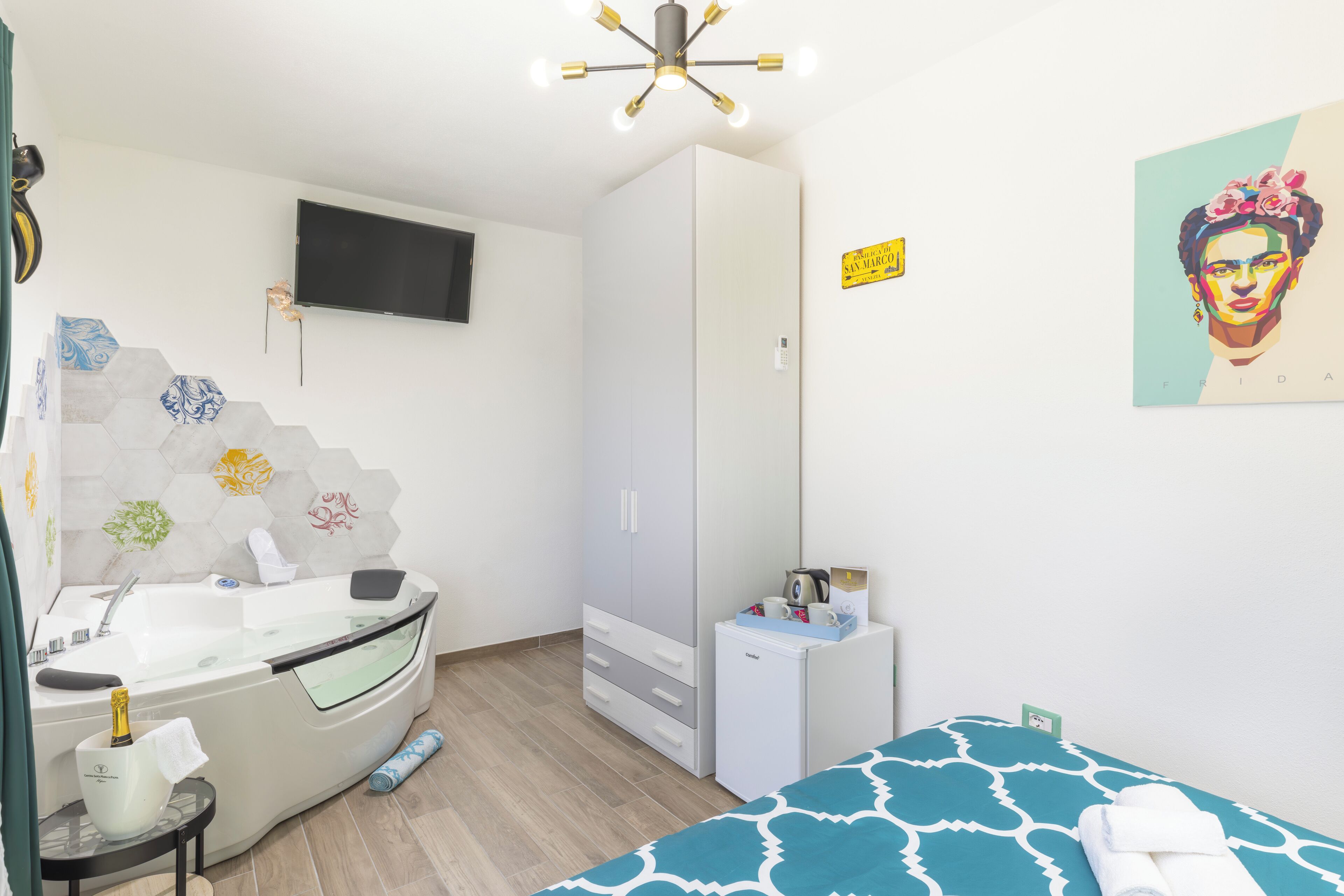 A modern bedroom with a bed, a vanity with a mirror, a small refrigerator, a TV mounted on the wall, and a ceiling light fixture.