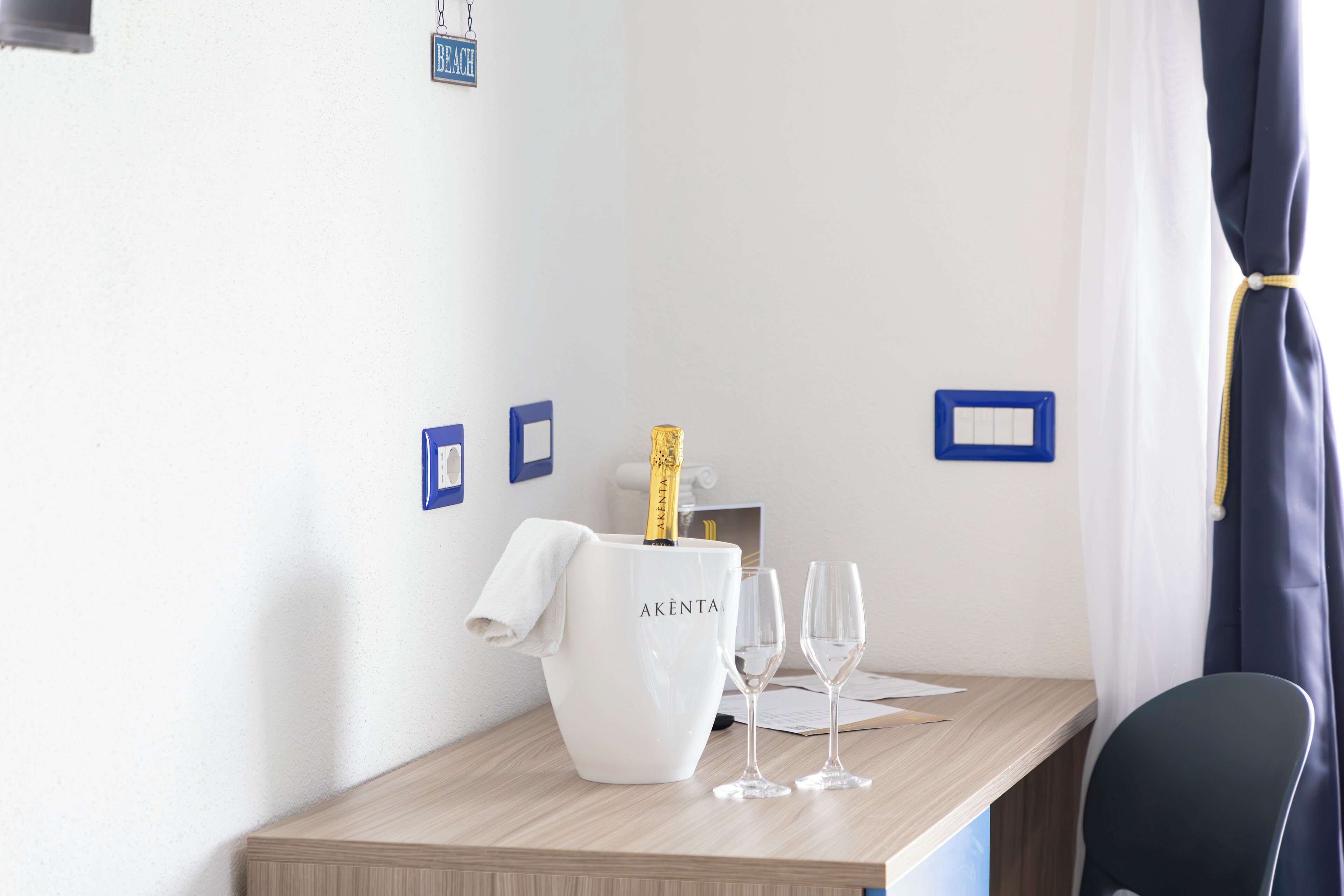 A hotel room with a wooden table, a white bucket labeled "AKENTA," two empty wine glasses, and a bottle of champagne.