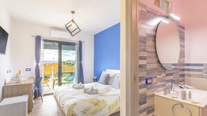 A hotel room with a bed, a desk, a chair, a small table, a bathroom with a mirror and sink, and a view of the outside. - Agua Dorada Retreat (Lula)