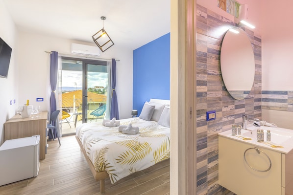 Habitación de hotel con cama, escritorio, silla, mesita, baño con espejo y lavamanos, y vista al exterior.