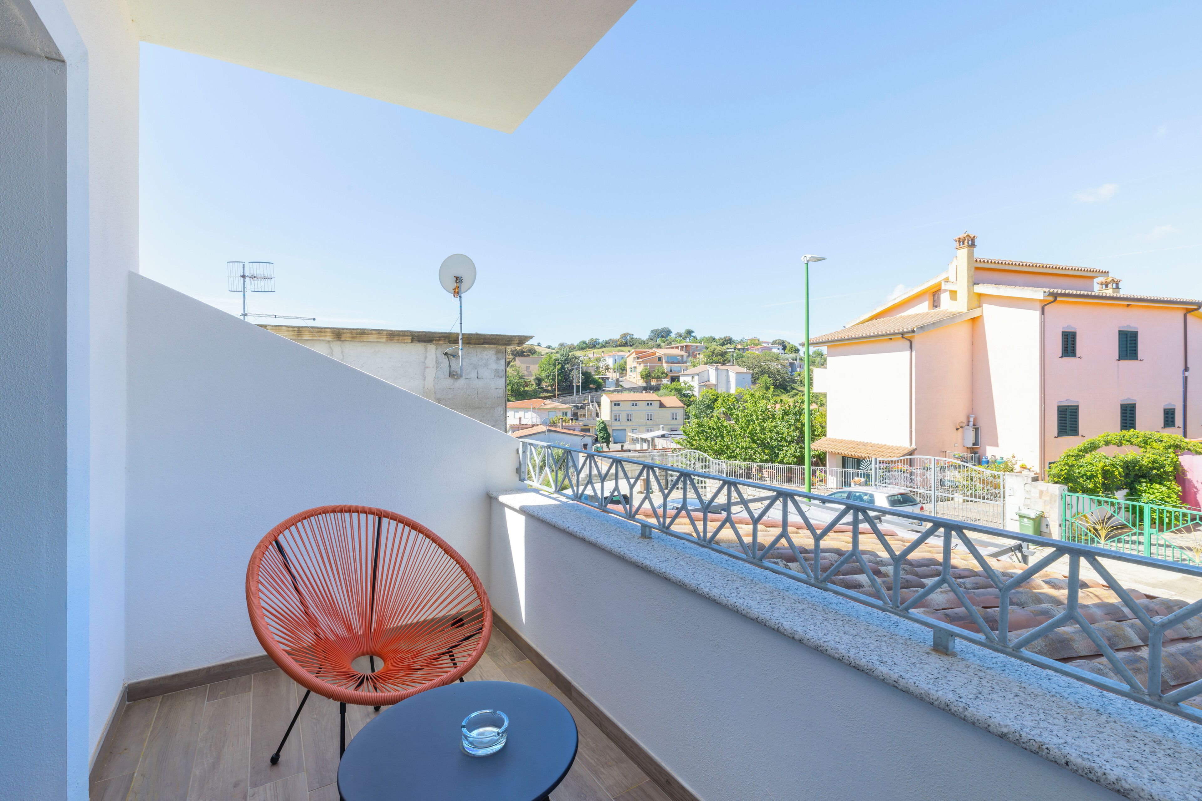 Un balcón con una mesa redonda y una silla roja, con vistas a una zona residencial con edificios y cielo despejado.