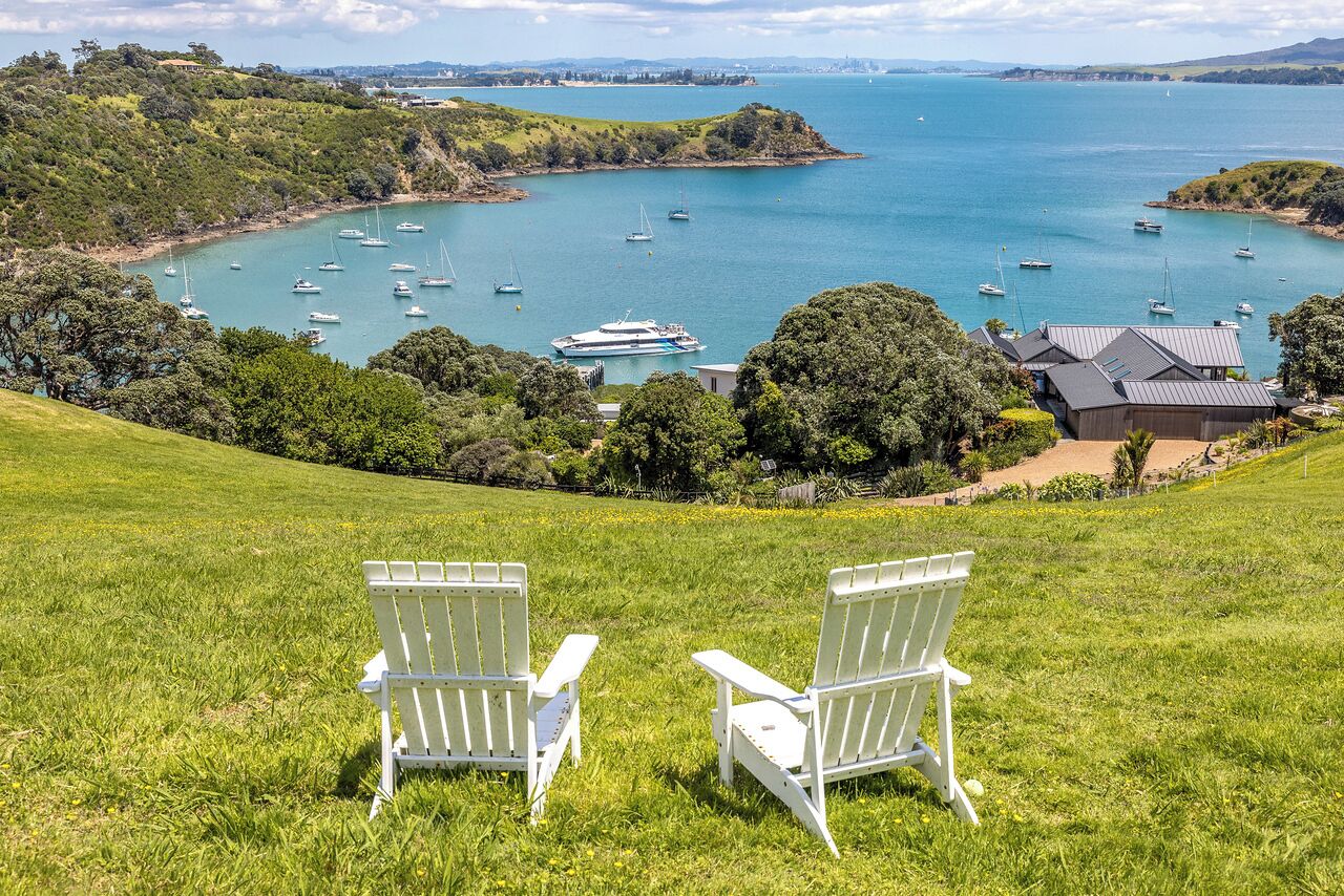 Dos sillas Adirondack blancas en una colina cubierta de césped con vista a una bahía con barcos.