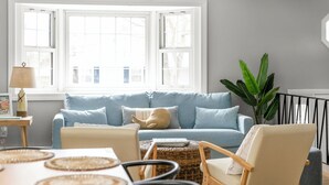 Una sala de estar con un sofá azul claro, una silla de madera, una mesita con individuales tejidos y una planta verde grande.