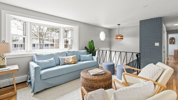 Una sala de estar moderna con un sofá azul claro, un reposapiés de mimbre y sillones blancos. Un ventanal ofrece vistas al exterior.
