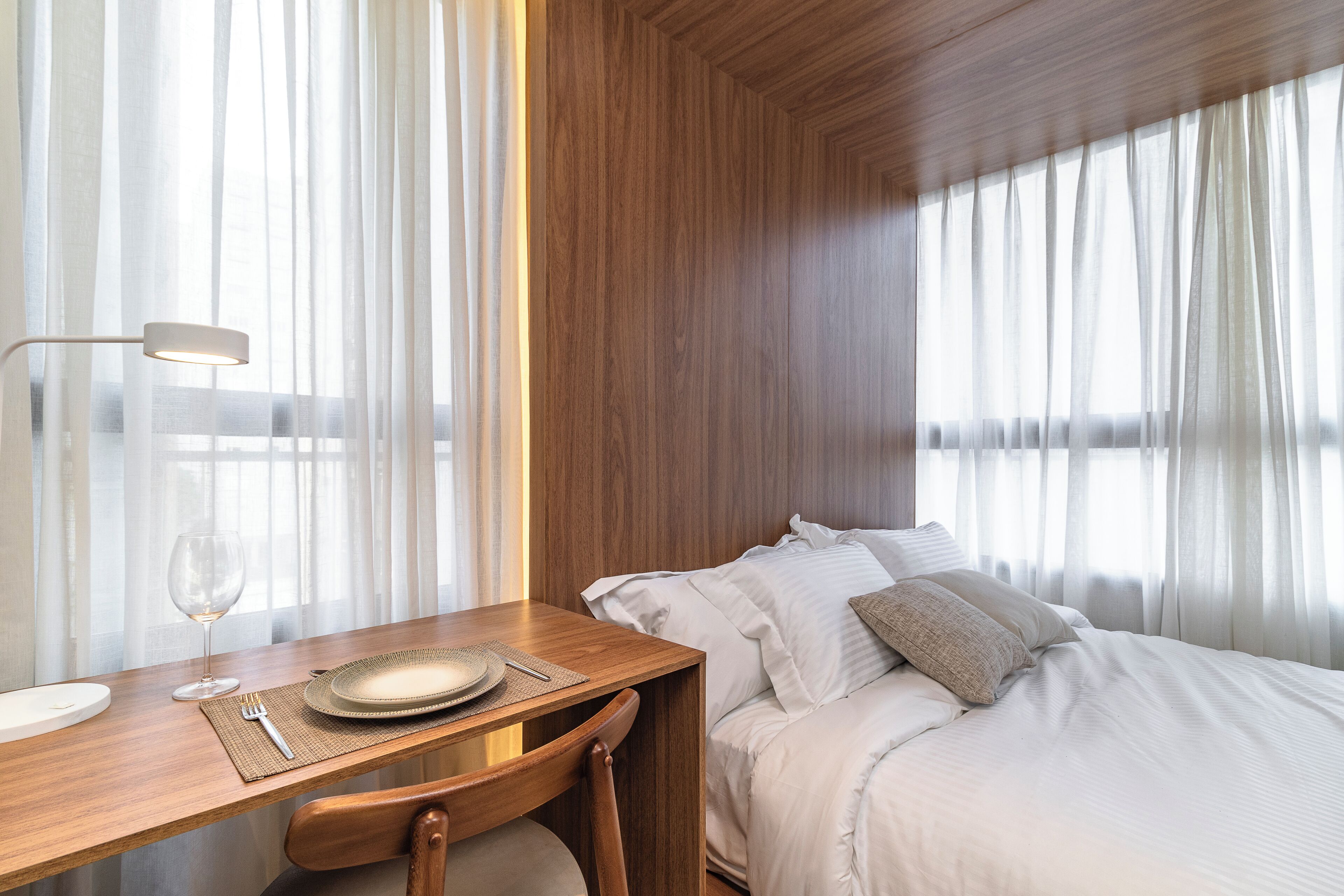 A hotel room with a wooden dining table, a glass wine glass, a plate, and a fork. There is a bed with white bedding and a pillow.
