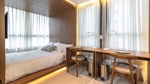 Una habitación de hotel moderna con una cama, un escritorio de madera y una silla. Hay cortinas en las ventanas, y se puede ver un cubo de basura.