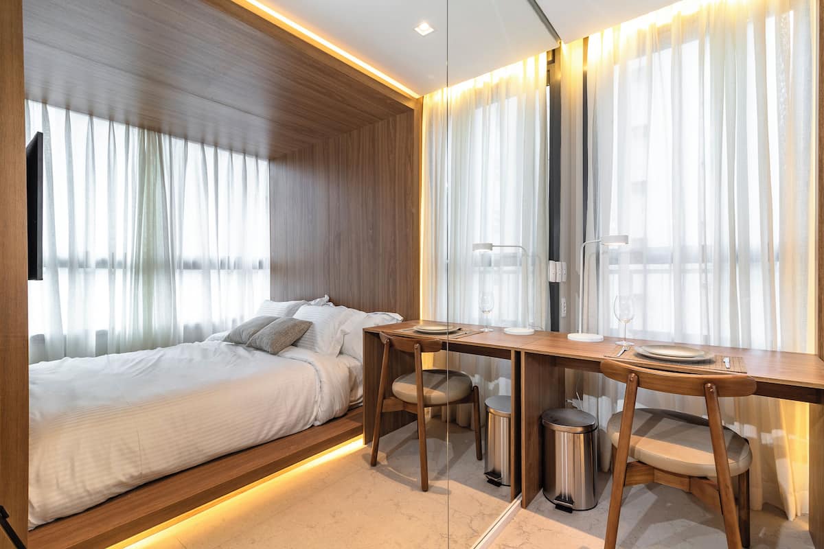 Une chambre d’hôtel moderne avec un lit, un bureau en bois et une chaise. Les fenêtres sont dotées de rideaux, et une poubelle est visible.