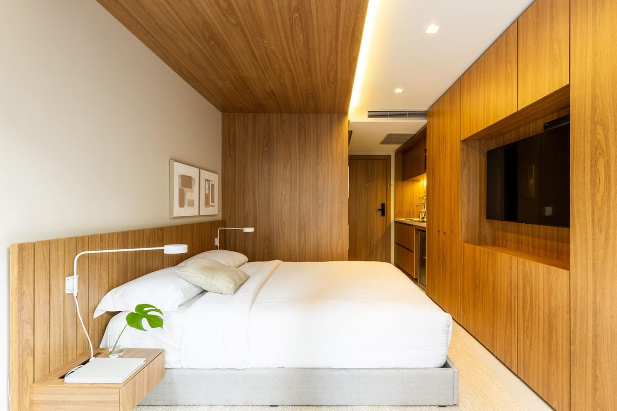 Une chambre moderne avec une tête de lit en bois, un lit avec une literie blanche, une table de chevet avec une lampe et un téléviseur fixé au mur.