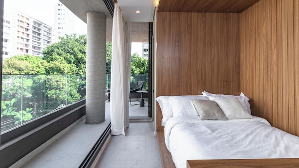 Un dormitorio moderno con un ventanal grande, una cama con ropa de cama blanca y vistas a un paisaje urbano y zonas verdes.