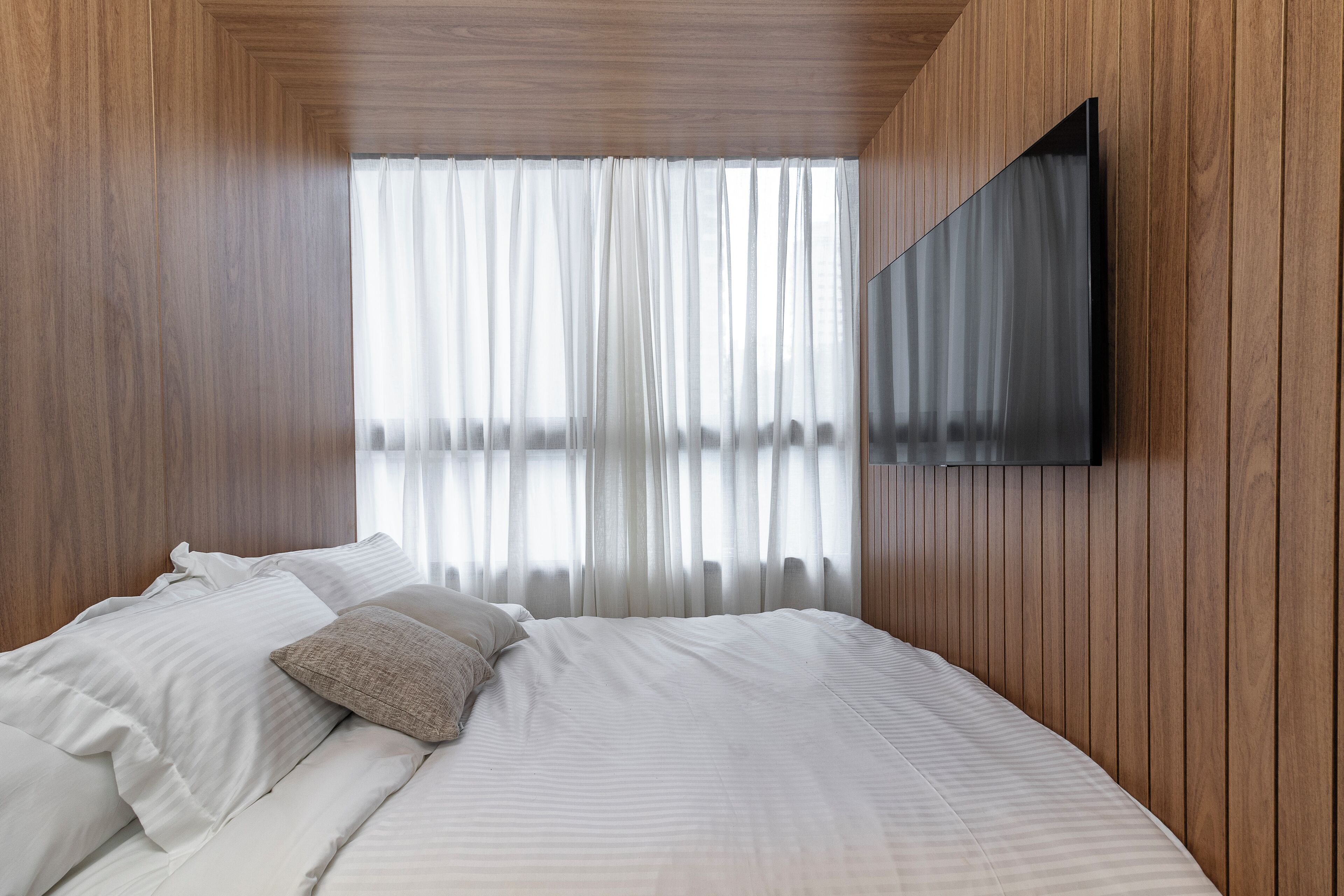 Une chambre à coucher avec un lit, du linge de lit blanc, un téléviseur fixé au mur et un lambris en bois.