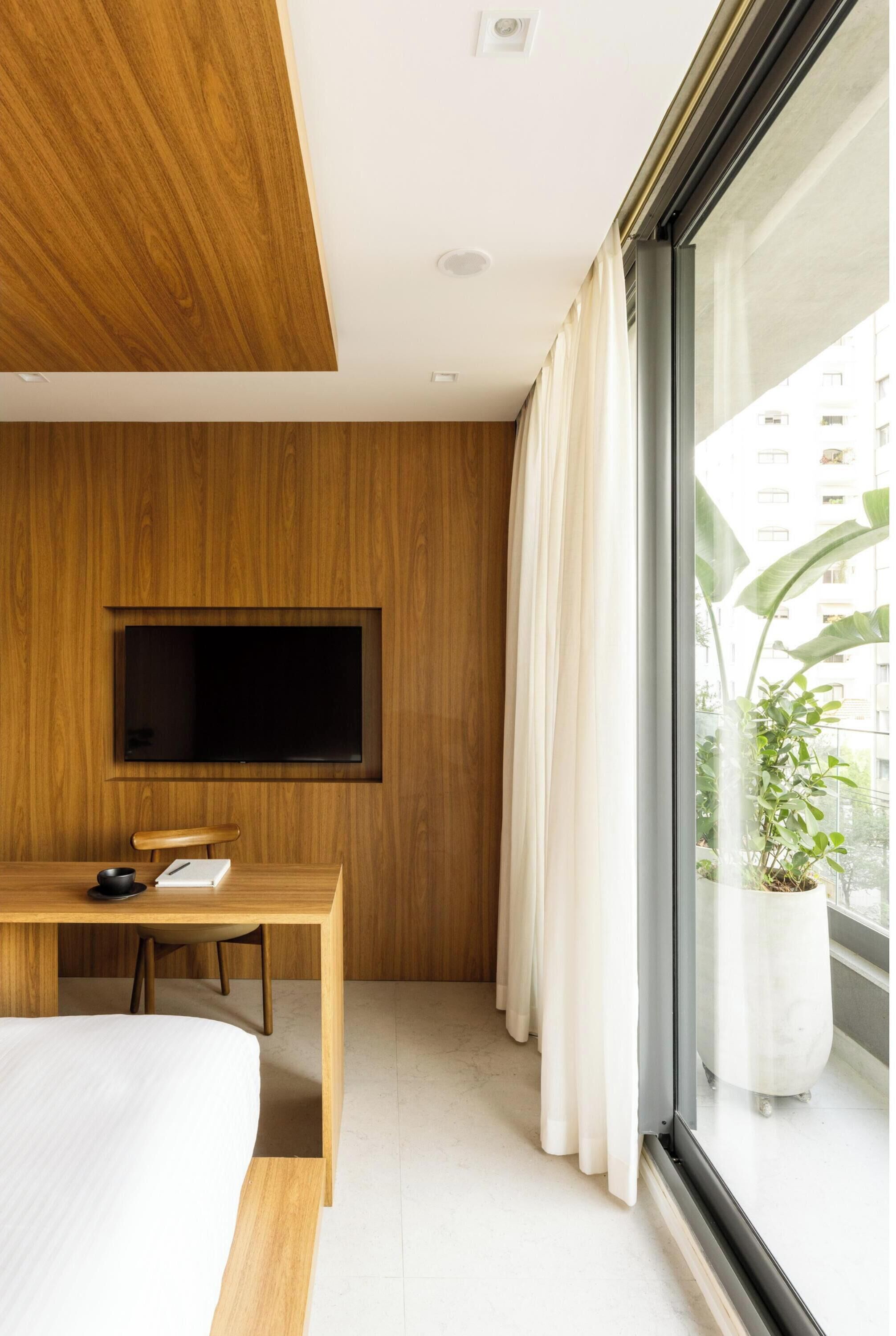 Une chambre d’hôtel moderne avec un mur revêtu de panneaux en bois, un téléviseur à écran plat fixé au-dessus d’un bureau en bois, un lit avec une literie blanche et une grande fenêtre avec des rideaux.