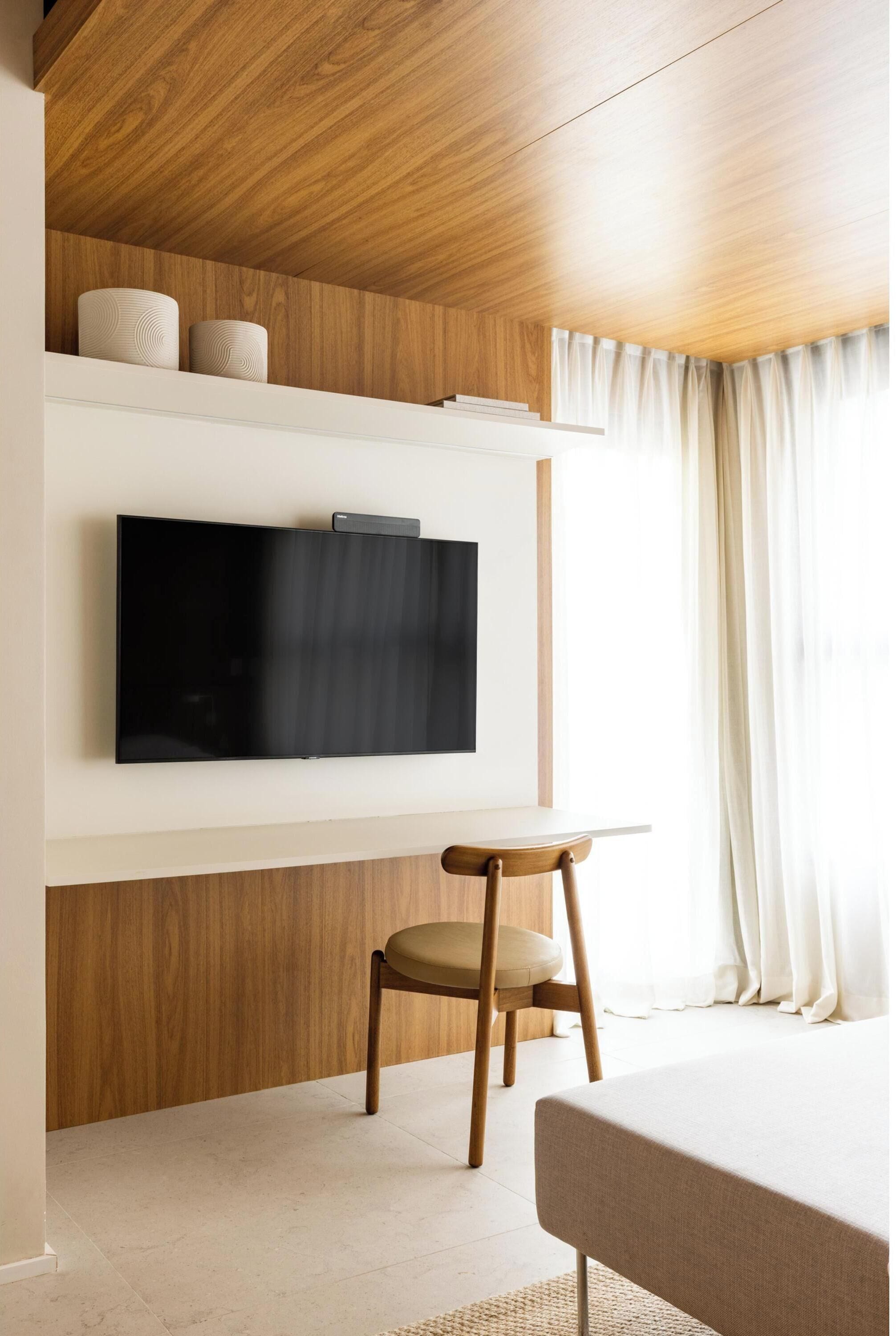 Una habitación moderna con un televisor de pantalla plana montado en una pared blanca, paneles de madera, una mesita redonda con una silla y una cama con cabecera blanca.