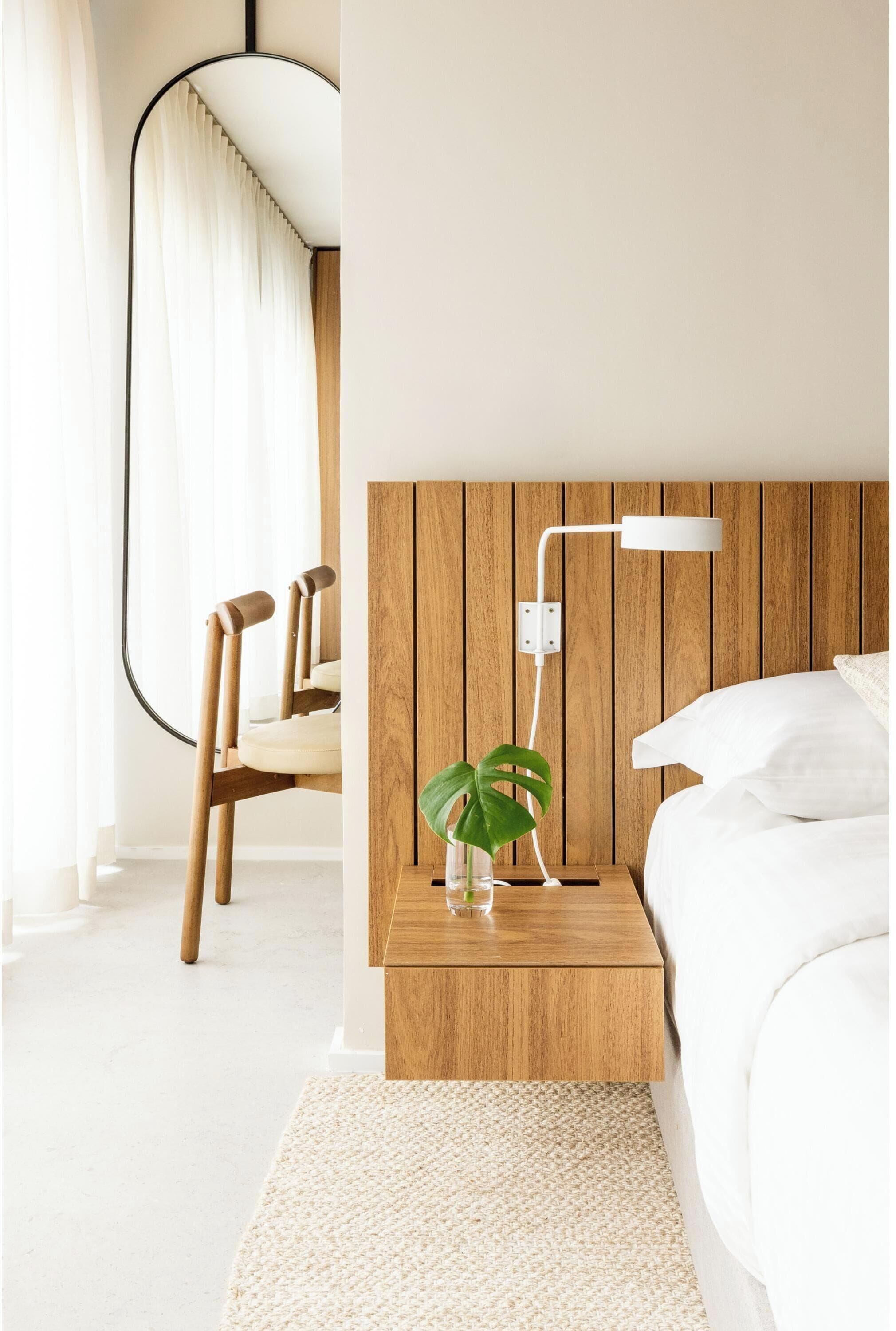A bedroom with a wooden headboard, a bedside table with a lamp, a plant, and a white bed.