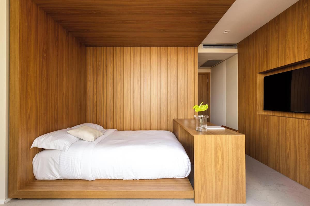 Une chambre moderne avec un mur lambrissé en bois, un lit recouvert de linge de lit blanc et une petite table de chevet en bois sur laquelle est posé un vase de fleurs.
