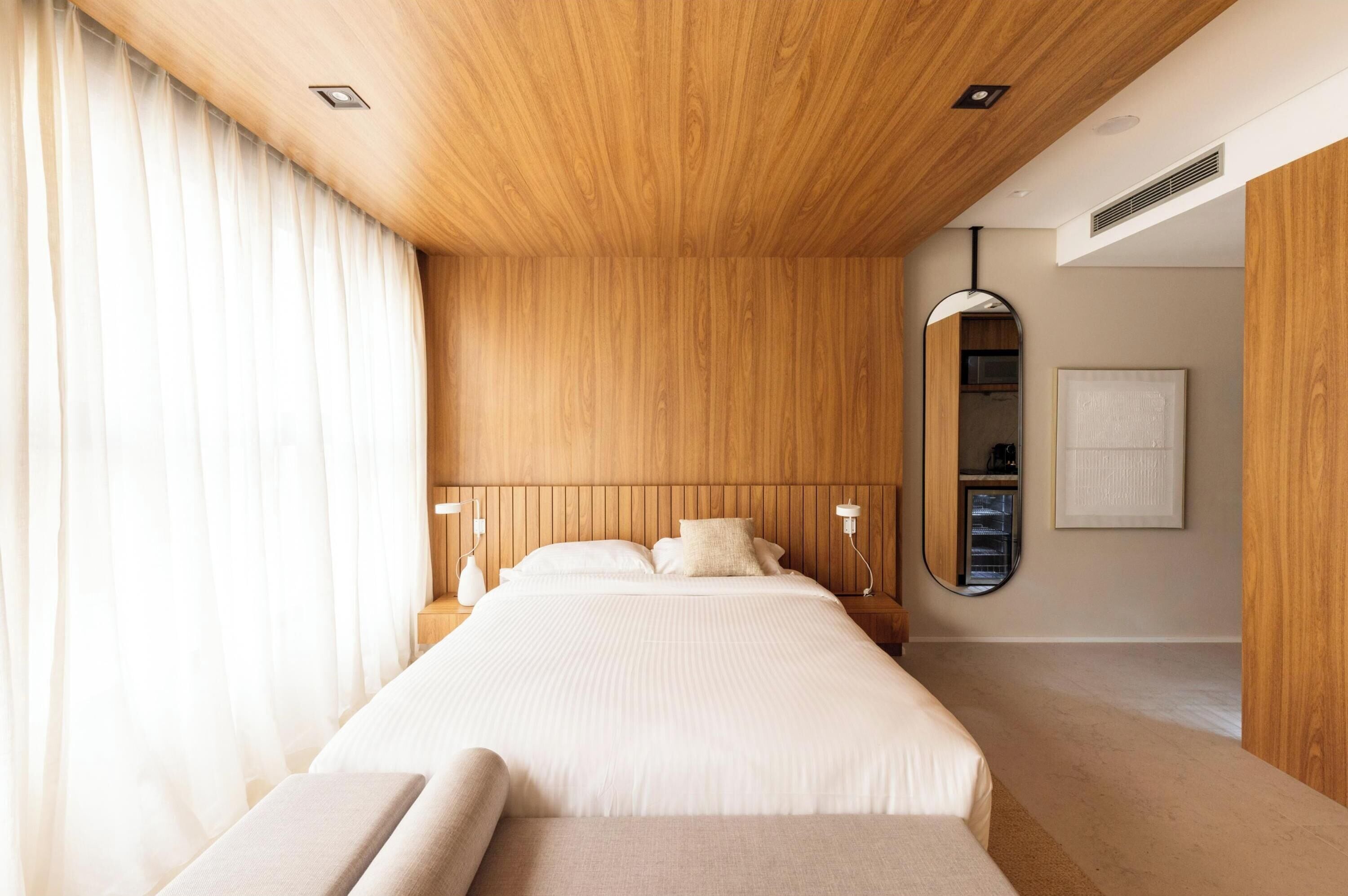 A modern bedroom with a wooden headboard, a large bed, bedside lamps, and a mirror on the wall.