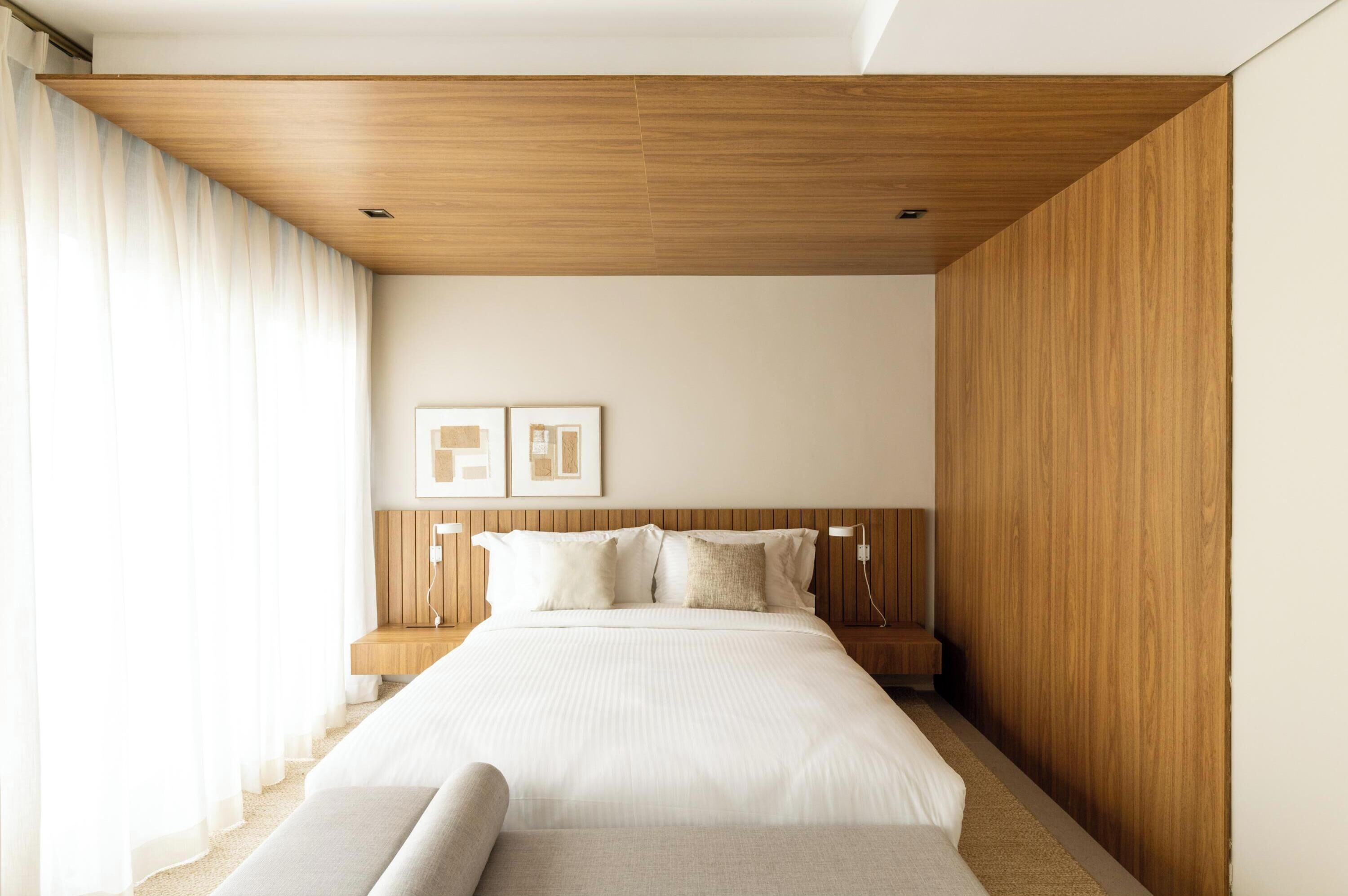 A modern bedroom with a wooden ceiling, a large bed with white bedding, a nightstand, and two framed pictures on the wall.