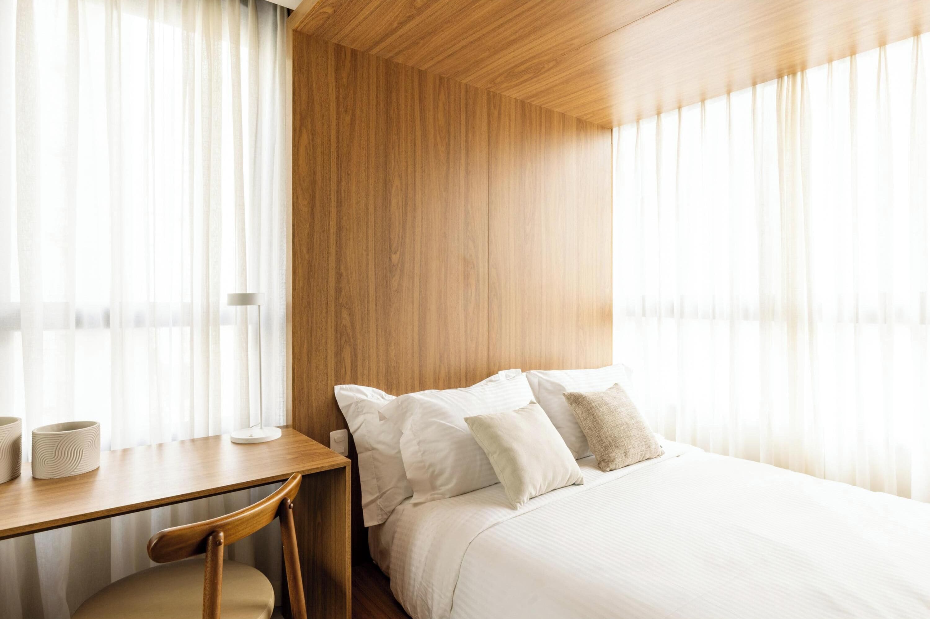 A hotel room with a wooden headboard, a bed with white bedding, a wooden desk with two white lamps, and a chair.