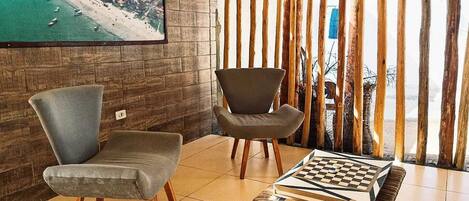 Une pièce avec un mur en bois, deux fauteuils gris rembourrés, une table basse en bois sur laquelle est posé un jeu d’échecs, et un tableau représentant une plage.