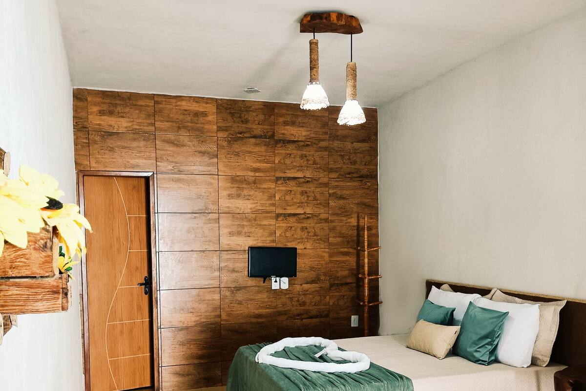 Un dormitorio con pared de paneles de madera, cama con ropa de cama verde y blanca, televisor montado en la pared y una mesa de madera con objetos decorativos.