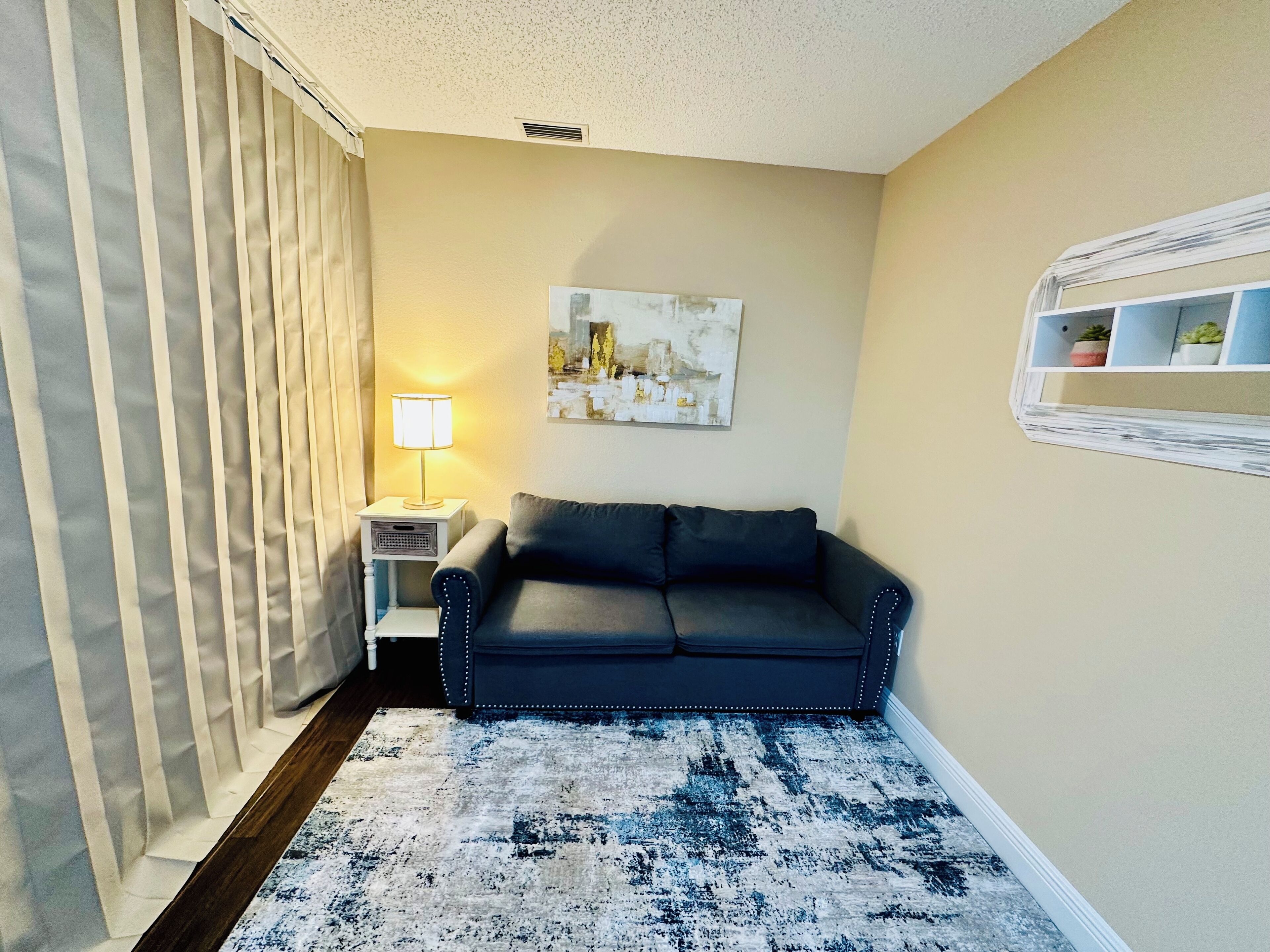 A small living room with a dark grey sofa, a white side table with a lamp, a patterned rug, a painting on the wall, and a white shelf with plants.