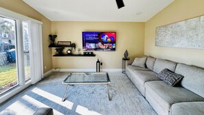 Un salon avec une table basse en verre, un canapé gris, un ventilateur de plafond et un téléviseur fixé au mur.