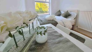 A bedroom with a bed, pillows, a bedside table with a vase of flowers, and a window with a view of trees. - Easy Rooms - London Luton Airport (Luton)