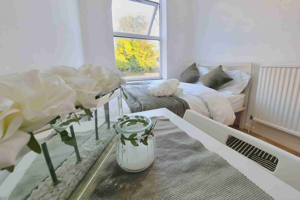 Ein Schlafzimmer mit Bett, Kissen, einem Nachttisch mit Blumen und einem Fenster mit Blick auf Bäume.