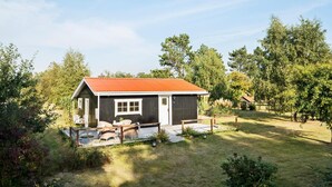 A small cabin with a red roof, a porch with outdoor furniture, and a grassy yard surrounded by trees.